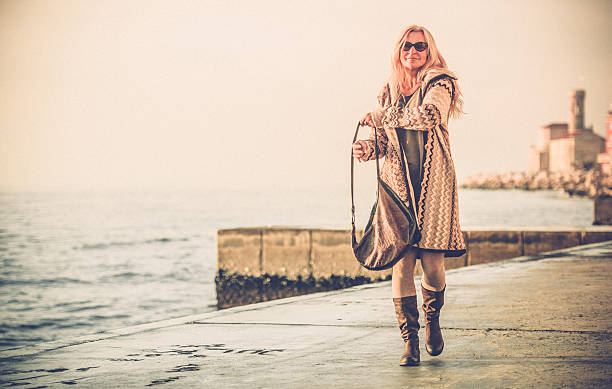 Attractive Mature Woman In Woolen Clothings Walking Attractive mature woman with long blond hair in woolen clothings in nature colors walking at seaside on a sunny winter day. Horizontal color image. Selective focus, back lit. Piran, Slovenia, Europe. older women short skirts stock pictures, royalty-free photos & images