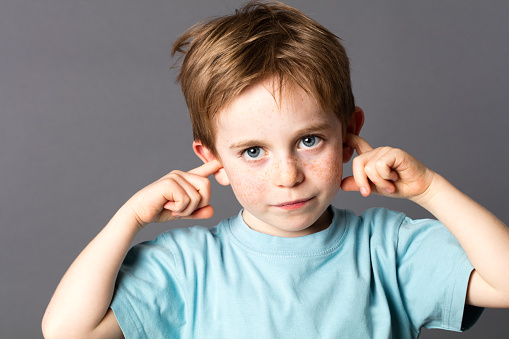sad little boy with blue eyes and freckles not willing to listen to domestic violence or parent relationship problems, covering his closed ears, grey background