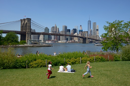 New York, USA - May 20, 2016: A view of the Brooklyn Bridge and the Manhattan skyline from Brooklyn Bridge Park. A couple is picnicking and children are playing during a warm and sunny day. Brooklyn Bridge Park is an 85-acre waterfront park on the Brooklyn side of the East River. It is visited by millions each year for it's view of the iconic Brooklyn Bridge and NYC skyline.