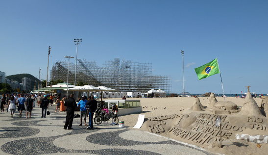Copacabana, Rio de Janeiro, Brazil - June 12, 2016: View of the symbolic sidewalk of Copacabana with people, a sand castle, a Brazilian flag, the beach and the structures of the future Olympic beach volleyball complex.