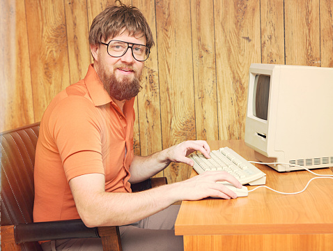 vintage inspired image of a nerdy man with with beard in a home office at his retro 1980's computer.  Wood paneling background
