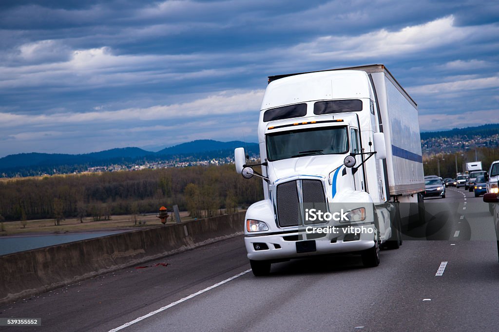 Big Rig American semi truck carring cargo on highway Elegant powerful semi truck with the windows on high cab for long distance moves along the highway with a load at dry van trailer. This rig is a true icon style on the American road. American Culture Stock Photo