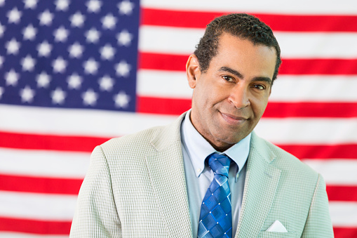 African American man standing in front of the American flag. He is wearing a suit and tie.