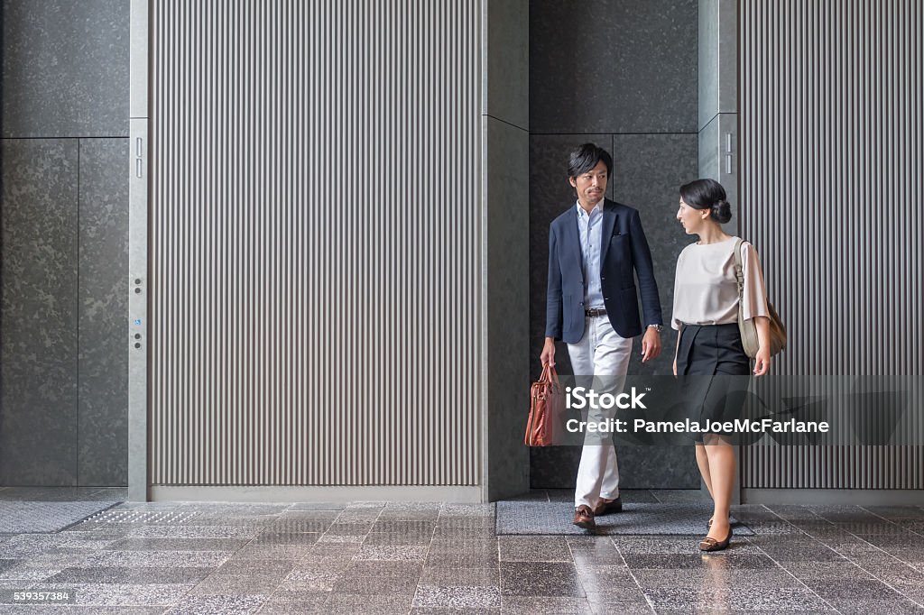 Japanese Businessman and Businesswoman Exiting Elevator in Modern Office Building Kyoto iStockalypse.  Asian man and woman having a discussion as they walk past an elevator in an office building lobby.  Kyoto, Japan. Walking Stock Photo