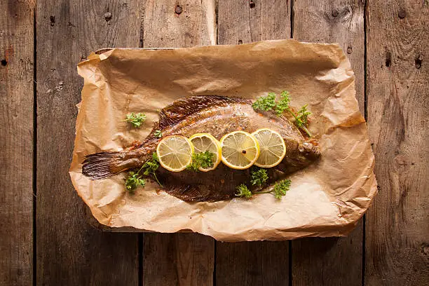 A whole baked Plaice covered in garnish on baking paper, onto of a rustic wooden background.