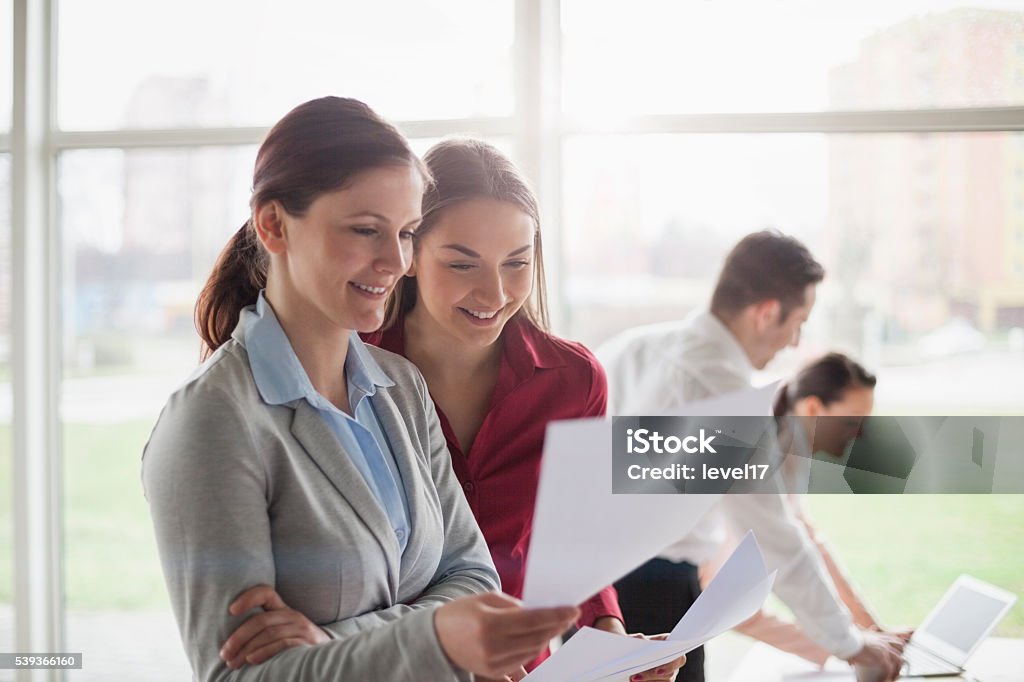Junge Geschäftsfrauen prüfen Dokumente mit Kollegen im Hintergrund - Lizenzfrei Anzug Stock-Foto