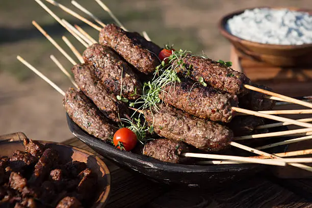 A bowl full of lamb koftas with tzatziki in the background, lit with sunlight in an outdoors picnic setting. 