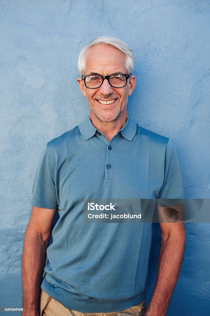Handsome mature man smiling at camera Portrait of handsome mature man with glasses looking at camera and smiling, he is leaning to against a blue wall. Mature Men Stock Photo