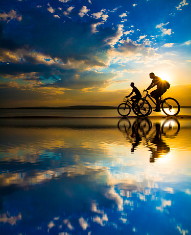 Empty Space. Sporty friends on bicycle on sunset. Silhouette couple cyclist go along shoreline coast. Sport in Nature background. Group of people two mountain biker in sunrise with reflection sun in water