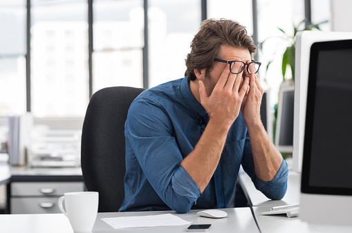 Portrait of an upset businessman at desk in office. Businessman being depressed by working in office. Young stressed business man feeling strain in eyes after working for long hours on computer.