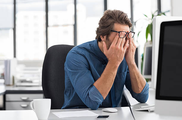 Stressed young businessman Portrait of an upset businessman at desk in office. Businessman being depressed by working in office. Young stressed business man feeling strain in eyes after working for long hours on computer. exhaustion stock pictures, royalty-free photos & images