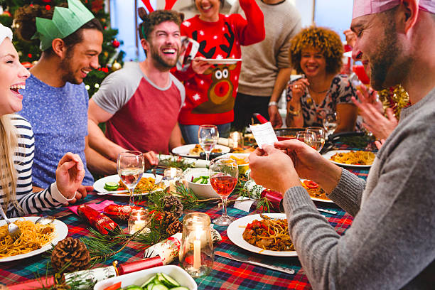 Christmas Cracker Group of friends laughing as they sit around a table eating and drinking at Christmas. One man is reading a joke from a christmas cracker. Party hats and christmas jumpers are worn. christmas cracker stock pictures, royalty-free photos & images