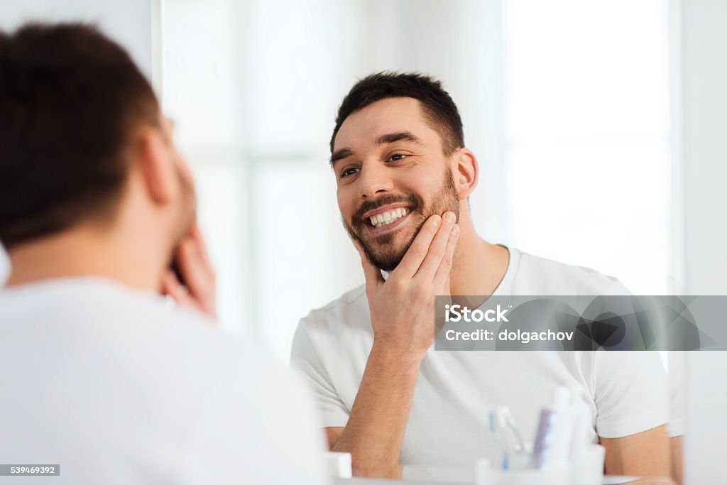 happy young man looking to mirror at home bathroom beauty, hygiene and people concept - smiling young man looking to mirror at home bathroom Men Stock Photo