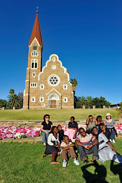 Windhoek, Namibia - September 19, 2009: Group of ,mainly African ,people sitting together on the lawn in front of the Lutheran Christuskirche ( Christ Church), a historical landmark in Windhoek,Namibia. They are laughing and having a good time whilst photographers are taking puctures of them. You can see the shadow of the photographers in front of them. Behind the people is a flowerbed with blooming azalea flowers.