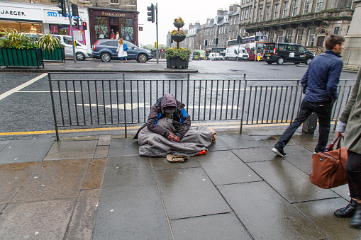 Edinburgh, UK: June 27, 2016: A beggar sits in the rain on a busy junction in Edinburgh. It is raining and he is being ignored by passersby. Begging is illegal under the Vagrancy Act of 1824. However it does not carry a jail sentence and is not well enforced in many cities.