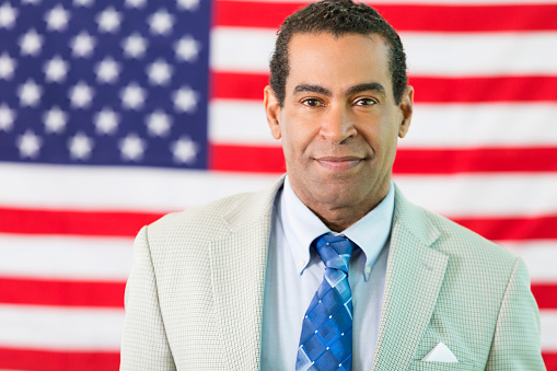 African American man standing in front of the American flag. He is wearing a suit and tie.