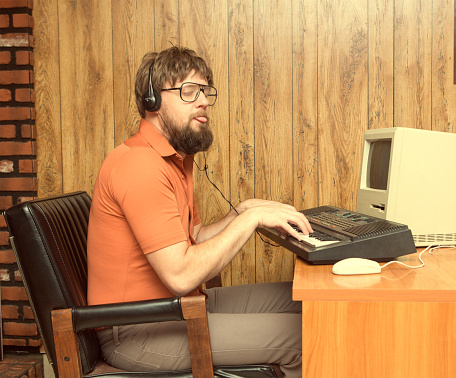 Nerdy 1980s themed photo of man playing a synthesizer keyboard with his retro computer recording music. Wearing Headphones