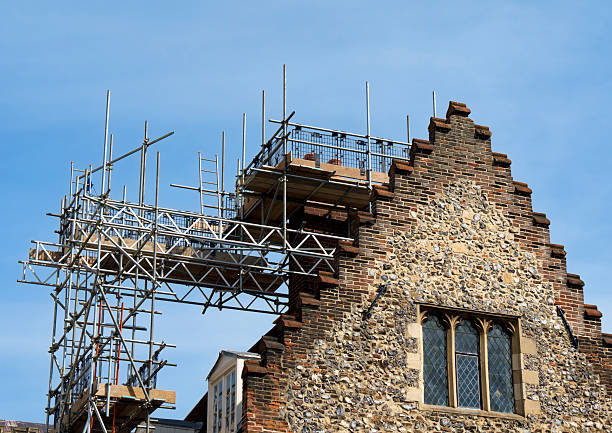 Scaffolding erected on an old building so that the construction workers can reach the chimneys and the roof. The building, in Norwich, Norfolk, eastern England, has a stepped gable and is constructed from brick and local flint.