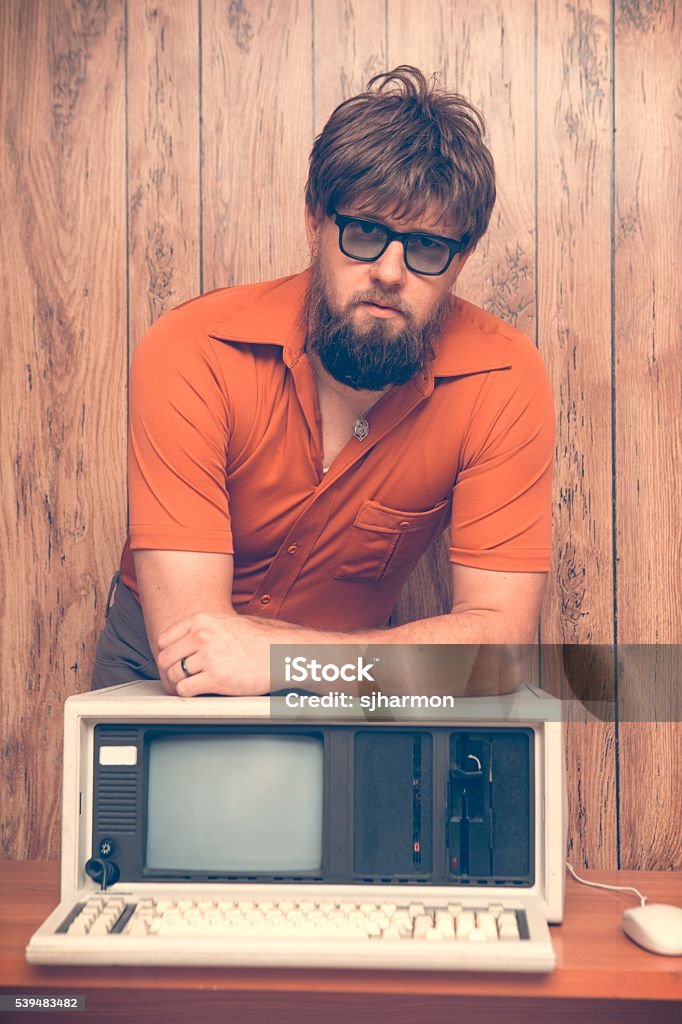 Vintage 1980's office worker with old computer Funny but serious looking retro 1980s business man leaning on his vintage computer. Wood panel background.  Man is wearing polyester orange shirt, glasses and has a neck beard. 1980-1989 Stock Photo