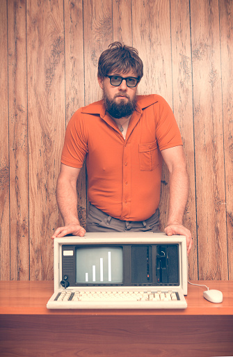 Funny but serious looking retro 1980s business man leaning on his vintage computer. Wood panel background.  Man is wearing polyester orange shirt, glasses and has a neck beard.