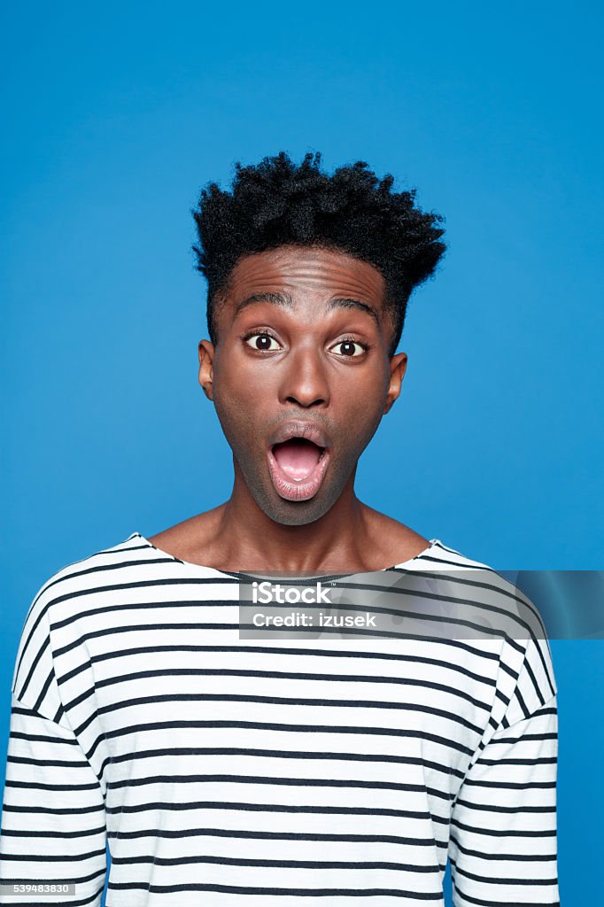 Surprised afro american young man Portrait of surprised afro american guy wearing striped long sleeved t-shirt, staring at camera with mouth open. Studio shot, blue background.  Adult Stock Photo