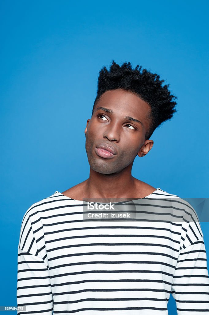 Pensive afro american young man Portrait of pensive afro american guy wearing striped long sleeved t-shirt, thinking, looking away. Studio shot, blue background.  Blue Background Stock Photo