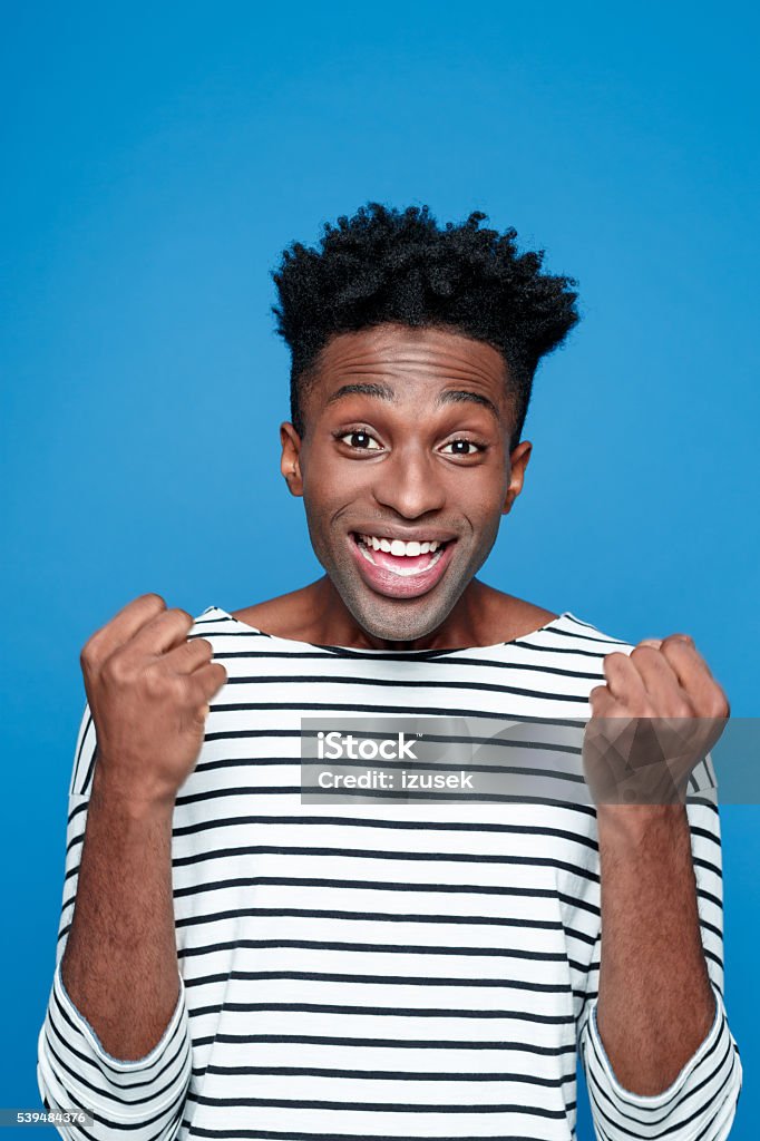 Excited afro american young man Portrait of happy afro american guy wearing striped long sleeved t-shirt, staring at camera with big smile, clenches his fists. Studio shot, blue background.  African Ethnicity Stock Photo