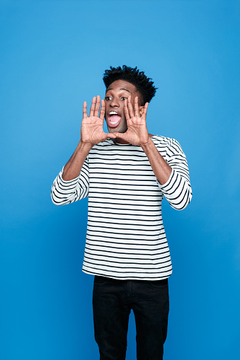 Portrait of excited afro american guy wearing striped long sleeved t-shirt and black trausers, scraming. Studio shot, blue background. 