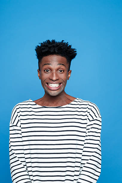 Excited afro american young man Portrait of happy afro american guy wearing striped long sleeved t-shirt, staring at camera. rolling eyes, raising eyebrows. Studio shot, blue background.  cheesy grin photos stock pictures, royalty-free photos & images
