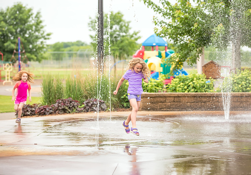 Two young girls (sisters) having fun running through the sprinklers at an outdour splash park on a cloudy summer day. The older sister is wearing a purple rash guard swim shirt and swim shorts with purple sandals. The younger sister is following her. She has curly red hair and is wearing a pink rash guard swim shirt and swim shorts.