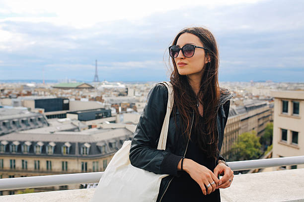 Vintage toned image of a young Parisian woman having a rest on the rooftop view in Paris, France, enjoying the view on a nice spring - early summer day. She is wearing a black leather jacket, and sunglasses, carrying a bag. Paris, lifestyle, street style fashion concepts.