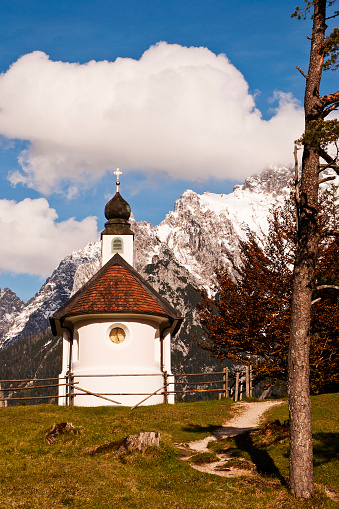 Vertical color photo of small chapel painted in pink and white near Mittenwald in front of majestic Karwendel mountain range under blue sky with clouds. Bavaria, Germany, May 2016