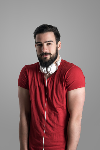 Young bearded casual man with headphones on neck posing with hands in pockets and shoulders raised up