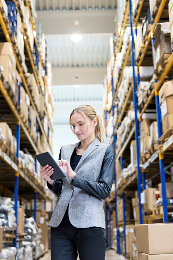 Vertical color image of confident young woman supervisor holding and touching digital tablet in warehouse. Young blond woman standing at distribution warehouse and wearing elegant suit. Industrial boss sincerely smiling and looking at camera. Logistic worker working in a large distribution warehouse. Large distribution storage in background with racks and shelfs full of packages, boxes, pallets and crates ready to be delivered. Logistics, freight, shipping, receiving.