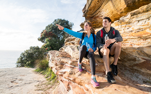 Happy couple hiking by the sea and pointing away at the view while sitting on a rock