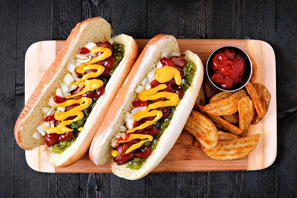 Hot dogs and potato wedges on wooden board, overhead view Two hot dogs fully loaded with toppings and potato wedges on wooden board, overhead view relish stock pictures, royalty-free photos & images