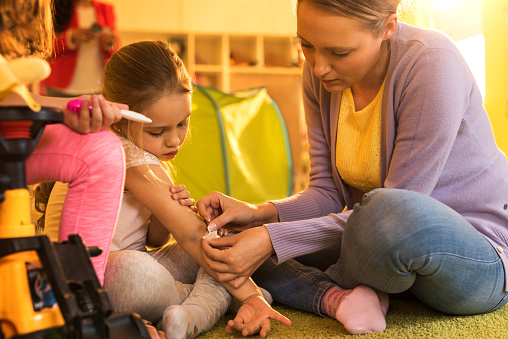 Teacher placing adhesive bandage on little girl's injured arm at preschool.