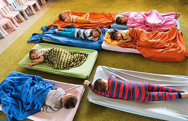 Above view of preschoolers in pajamas sleeping at kindergarten. High angle view of group of tired kids in pajamas resting on beds at kindergarten. napping stock pictures, royalty-free photos & images