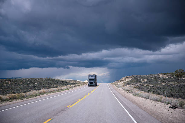 Dark grey semi-truck on highway and storming sky Dark gray semi truck with trailer approaching in the opposite lane level long road on the background of a stormy sky Nevada storming stock pictures, royalty-free photos & images