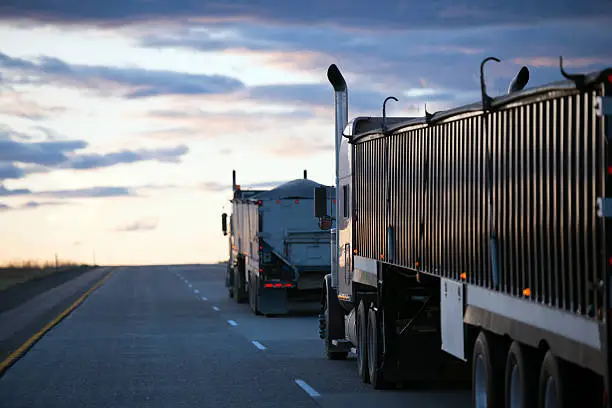 Several semi trucks with bulk cargo trailers move by convoy on the road towards of the evening horizon