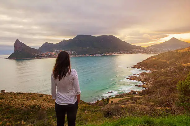 A tourist stands at a lookout point enjoying a beautiful view of the mountains and bay of Hout Bay, Cape Town
