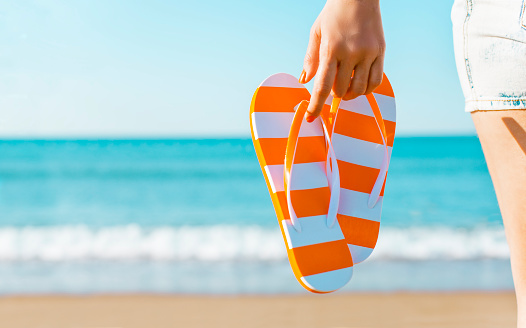 Woman's hand holding a pair of orange sandals against the coastline