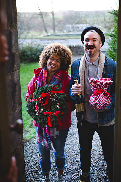 Christmas Visitors A happy young couple smile as they stand on the doors step visiting a friend. Both are wrapped up in warm clothing and hold gift, wreath and a bottle of champagne. front stoop photos stock pictures, royalty-free photos & images