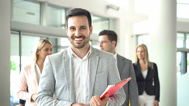 Team leadership. Closeup of handsome assertive businessman standing in front of his colleagues. He's smiling and looking at camera, wearing fray smart casual suit and holding a brochure. assertiveness stock pictures, royalty-free photos & images