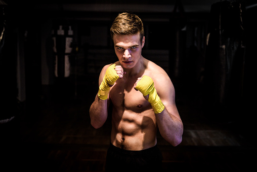 Street fighter mma boxer with bandages ready for fight club. Athletic ripped young man with big abs and wraps in a gym after a kickbox workout is looking at the camera.