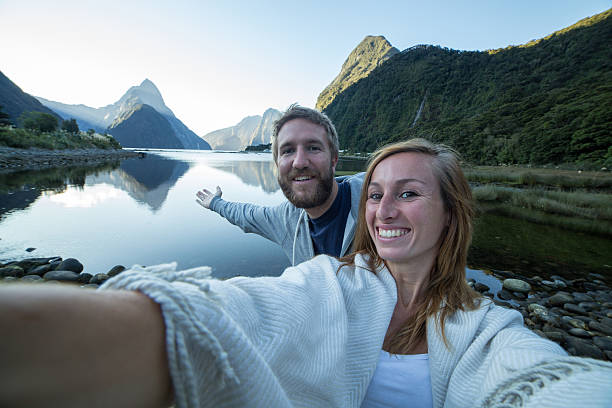 Cheering couple take a selfie portrait at Milford Sound, NZ Cheering young couple take a selfie portrait in front of the Mitre peak in Milford sound, New Zealand. mitre peak stock pictures, royalty-free photos & images
