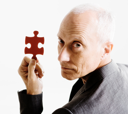 Silver-haired man looks over his shoulder at camera, holding up a single red jigsaw puzzle piece seriously.