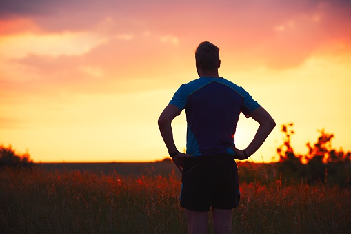Silhouette of runner. Outdoor cross-country running. Pensive young man is taking rest after running in the nature during golden sunset.