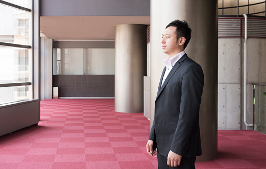 This is a horizontal, color photograph of one Japanese businessman dressed in a suit. He is standing in the interior of a large atrium with floor to ceiling windows in a modern Japanese office building. Lost in thought, he looks out towards the window with a serious look on his face. Photographed with a Nikon D800 DSLR camera in Kyoto, Japan.