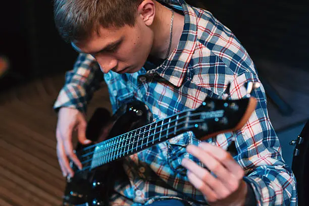 Musician tuning strings on his bass-guitar. Front view on bass player spinning guitar tuners. Closeup on young guy with bass guitar